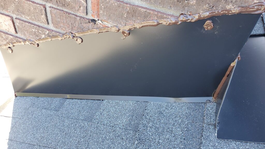 Here is a picture of the side of a red brick chimney with new black flashing along the side with brown sealant on it. 